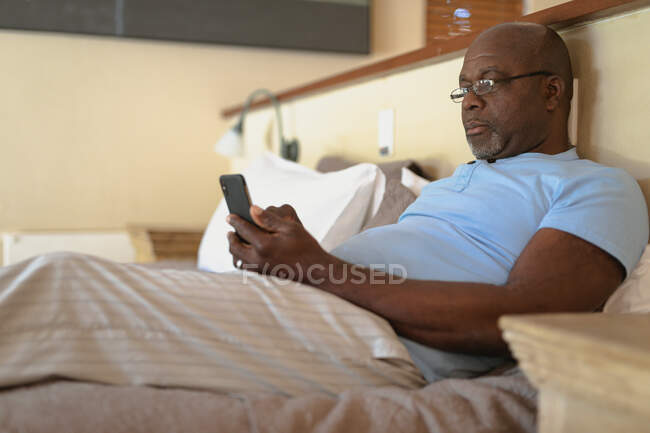 Ein älterer afrikanisch-amerikanischer Mann legt sich ins Bett und benutzt ein Smartphone. Lebensstil im Ruhestand, Zeit allein zu Hause verbringen. — Stockfoto