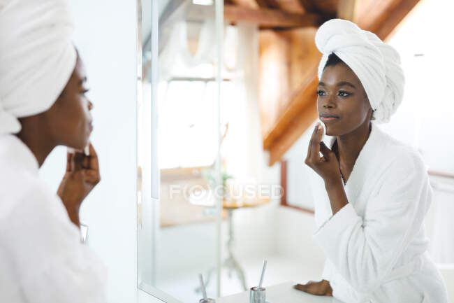 Femme afro-américaine souriante dans la salle de bain, visage nettoyant avec du coton pour les soins de la peau. mode de vie domestique, profiter de loisirs d'auto-soins à la maison. — Photo de stock