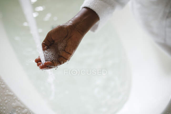 Gros plan de la main de la femme afro-américaine dans la salle de bain en cours d'exécution d'une baignoire. mode de vie domestique, profiter de loisirs d'auto-soins à la maison. — Photo de stock