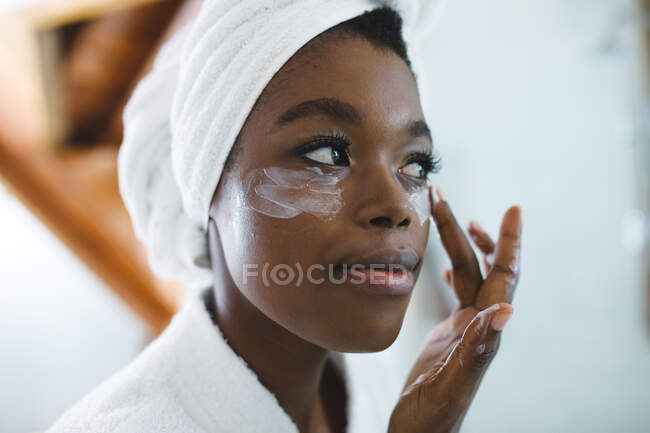 Mujer afroamericana sonriente en el baño aplicando crema facial para el cuidado de la piel. estilo de vida doméstico, disfrutando del tiempo libre de autocuidado en casa. - foto de stock