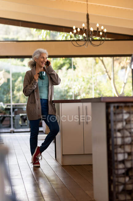 Старшая белая женщина на кухне использует смартфон и пьет кофе. пенсионного образа жизни, проводить время в одиночестве на дому. — стоковое фото