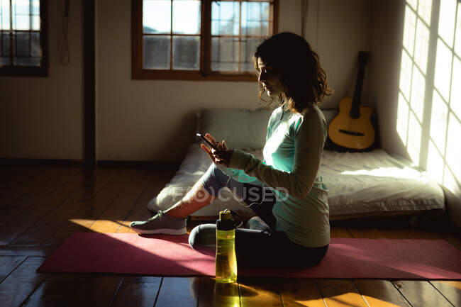 Femme de race mixte pratiquant le yoga, en utilisant un smartphone dans le salon ensoleillé. mode de vie sain, profiter de loisirs à la maison. — Photo de stock
