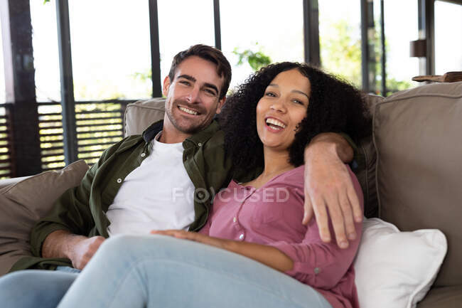 Portrait of happy diverse couple sitting on sofa in living room embracing and smiling. spending time off at home in modern apartment. — Stock Photo