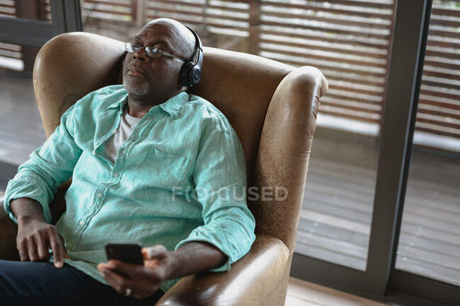 Détente homme afro-américain sénior assis et en utilisant des écouteurs dans le salon moderne. mode de vie à la retraite, passer du temps seul à la maison. — Photo de stock
