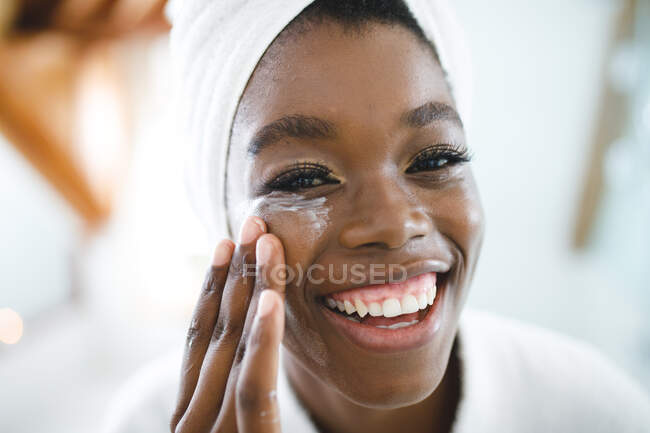Portrait de femme afro-américaine souriante dans la salle de bain appliquant de la crème visage pour les soins de la peau. mode de vie domestique, profiter de loisirs d'auto-soins à la maison. — Photo de stock