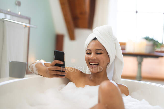 Laughing mixed race woman in bathroom having a bath and using smartphone. domestic lifestyle, enjoying self care leisure time at home. — Stock Photo
