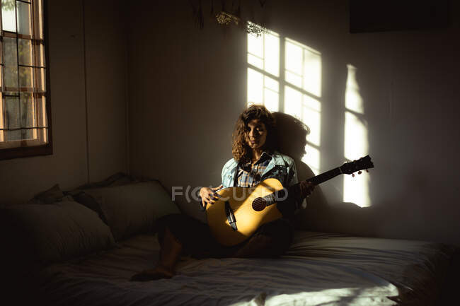 Mujer de raza mixta tocando la guitarra en el dormitorio soleado. estilo de vida saludable, disfrutando del tiempo libre en casa. - foto de stock