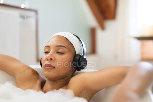 Смешанная женщина расы в ванной комнате расслабляется в ванне в наушниках, с закрытыми глазами. домашний образ жизни, наслаждаясь отдыхом на дому. — стоковое фото