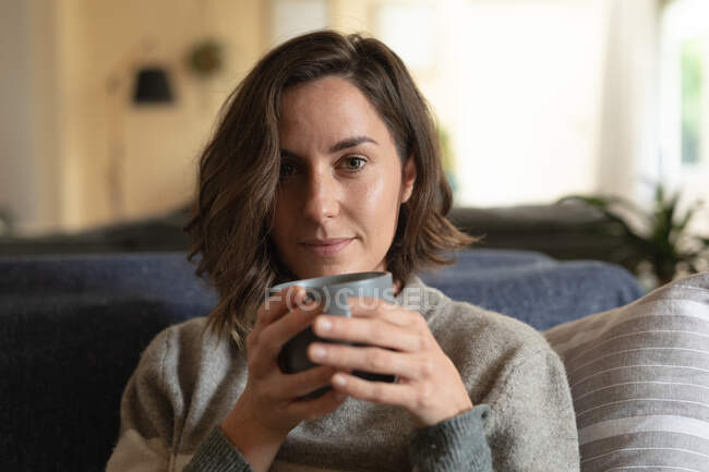 Portrait de femme caucasienne souriante dans le salon assis sur un canapé et buvant du café. mode de vie domestique, profiter du temps libre à la maison. — Photo de stock