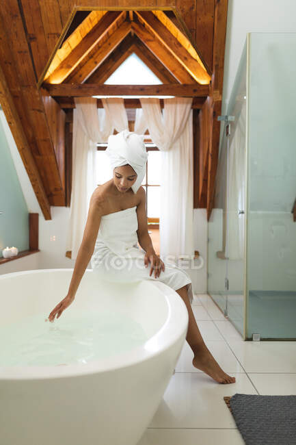 Femme de race mixte dans la salle de bain courir une baignoire, assis sur le bord de la baignoire. mode de vie domestique, profiter de loisirs d'auto-soins à la maison. — Photo de stock