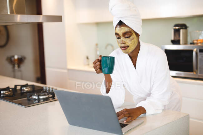 Femme afro-américaine souriante avec masque de beauté, dans la cuisine en utilisant un ordinateur portable et en buvant du café. mode de vie domestique, profiter de loisirs d'auto-soins à la maison. — Photo de stock