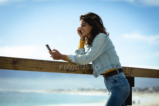 Femme de race mixte utilisant smartphone le jour ensoleillé au bord de la mer. mode de vie sain, profiter du temps libre à l'extérieur. — Photo de stock