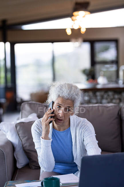 Donna caucasica anziana in soggiorno seduta sul divano, utilizzando smartphone e laptop. stile di vita di pensione, trascorrere del tempo da solo a casa. — Foto stock