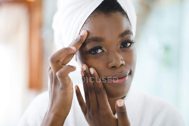 Portrait de femme afro-américaine souriante dans la salle de bain touchant son visage avant le traitement de beauté. mode de vie domestique, profiter de loisirs d'auto-soins à la maison. — Photo de stock