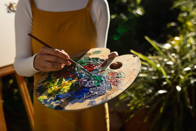 Mujer caucásica en jardín soleado, mezclando pintura en la paleta. estilo de vida doméstico, disfrutando del tiempo libre en casa. - foto de stock
