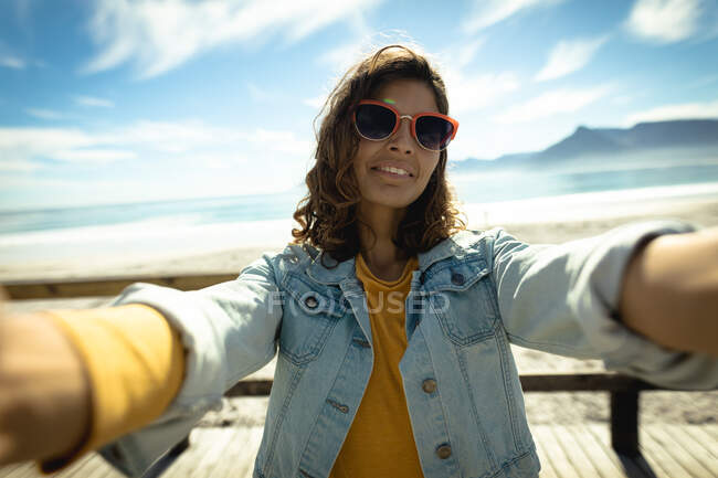 Mujer de raza mixta tomando selfie con smartphone en un día soleado junto al mar. estilo de vida saludable, disfrutar del tiempo libre al aire libre. - foto de stock