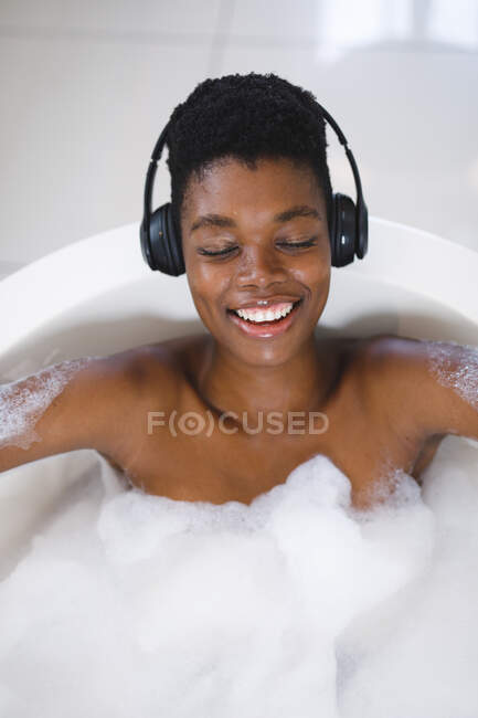 Femme afro-américaine souriante se détendre dans le bain avec les yeux fermés en écoutant de la musique sur écouteurs. — Photo de stock