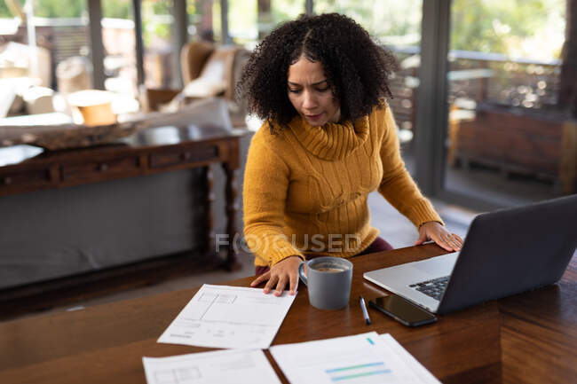 Frau mit gemischter Rasse sitzt am Tisch und arbeitet mit Laptop. Arbeiten zu Hause in moderner Wohnung. — Stockfoto