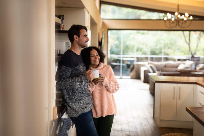 Feliz pareja diversa en la cocina abrazando beber café y sonreír. pasar tiempo libre en casa en apartamento moderno. - foto de stock