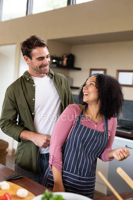Heureux couple diversifié dans la cuisine préparant la nourriture ensemble en se regardant et en souriant. passer du temps à la maison dans un appartement moderne. — Photo de stock