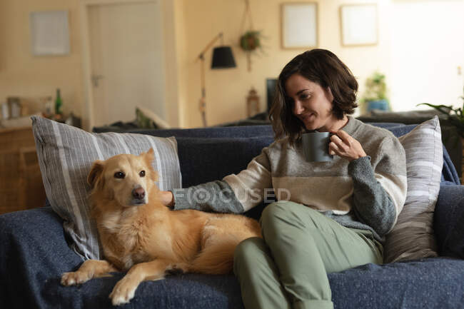 Mujer caucásica sonriente en la sala de estar sentada en el sofá acariciando a su perro mascota y tomando café. estilo de vida doméstico, disfrutando del tiempo libre en casa. - foto de stock