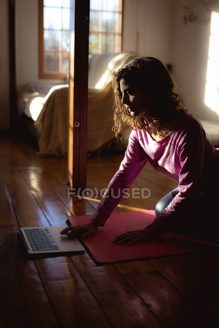 Mujer de raza mixta practicando yoga, utilizando portátil en la sala de estar soleada. estilo de vida saludable, disfrutando del tiempo libre en casa. - foto de stock