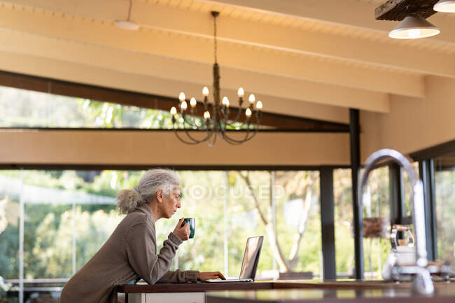 Mulher caucasiana sênior na cozinha usando laptop e beber café. estilo de vida aposentadoria, passar o tempo sozinho em casa. — Fotografia de Stock