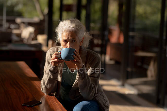 Mujer caucásica mayor en la cocina sentado y bebiendo café. estilo de vida de jubilación, pasar tiempo solo en casa. - foto de stock