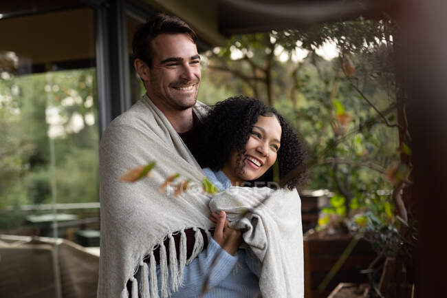 Счастливая, разнообразная пара, покрытая одеялом на балконе, обнимающая и улыбающаяся. проводить свободное время дома. — стоковое фото