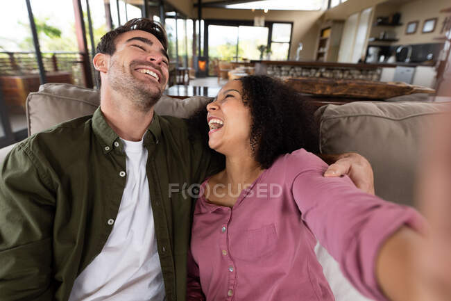 Щаслива різноманітна пара сидить на дивані у вітальні, приймаючи селфі та посміхаючись. вихідні вдома в сучасній квартирі . — стокове фото