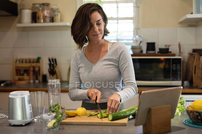 Белая женщина на кухне, готовит лечебный напиток, режет овощи, использует планшет. домашний образ жизни, наслаждаясь отдыхом дома. — стоковое фото