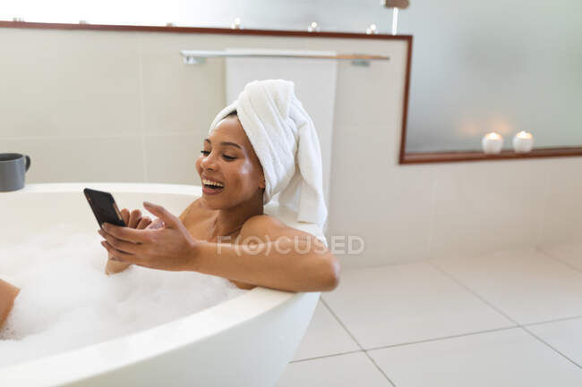 Riendo mujer de raza mixta en el baño con bañera y el uso de un teléfono inteligente. estilo de vida doméstico, disfrutando del tiempo libre de autocuidado en casa. - foto de stock