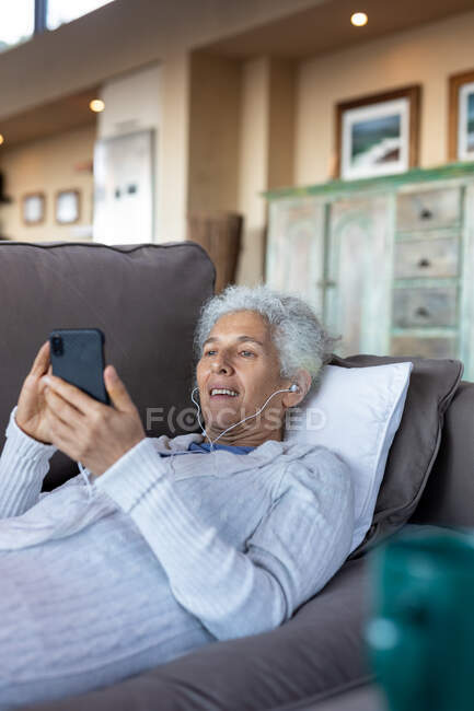 Happy senior caucasian woman laying and using smartphone in the modern living room. retirement lifestyle, spending time alone at home. — Stock Photo