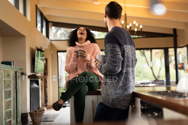 Heureux couple diversifié dans la cuisine assis sur le comptoir de boire du café et de parler. passer du temps à la maison dans un appartement moderne. — Photo de stock