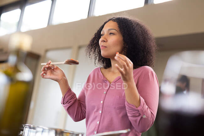 Feliz mujer de raza mixta en la cocina preparando comida y comiendo. pasar tiempo libre en casa en apartamento moderno. - foto de stock