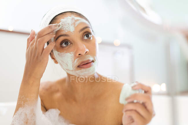 Gemischte Rassenfrau im Badezimmer, die eine Badewanne hat und eine Gesichtsmaske aufträgt. häuslicher Lebensstil, selbstgepflegte Freizeit zu Hause genießen. — Stockfoto