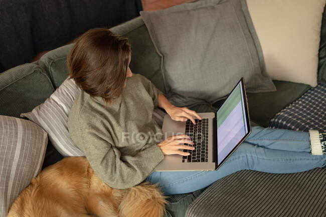 Кавказька жінка у вітальні, сидячи на дивані зі своїм собакою, за допомогою ноутбука. Домашнє життя, дозвілля вдома. — стокове фото