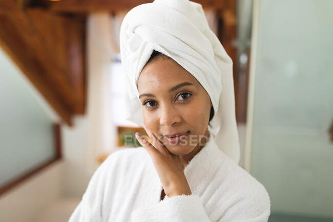Портрет усмішки змішаної раси жінка у ванній кімнаті торкається обличчя після зволоження, дивлячись на камеру. — стокове фото