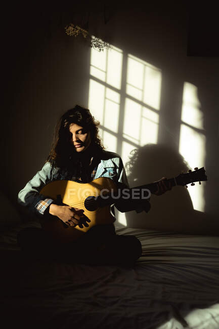 Mujer de raza mixta tocando la guitarra en el dormitorio soleado. estilo de vida saludable, disfrutando del tiempo libre en casa. - foto de stock