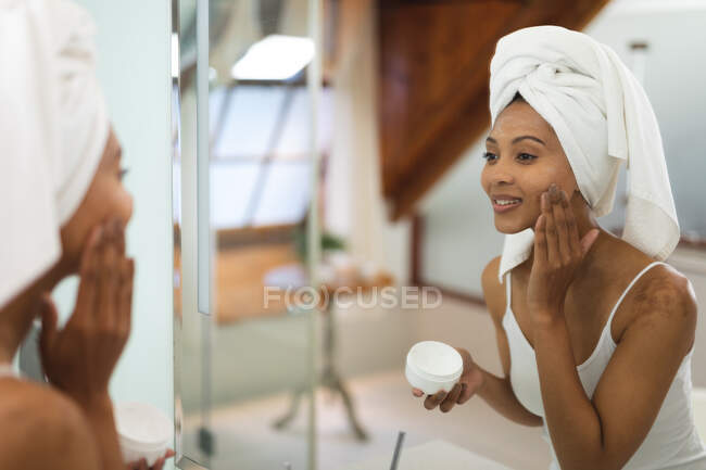 Mujer de raza mixta en baño aplicando crema facial para el cuidado de la piel, mirándose en espejo. estilo de vida doméstico, disfrutando del tiempo libre de autocuidado en casa. - foto de stock