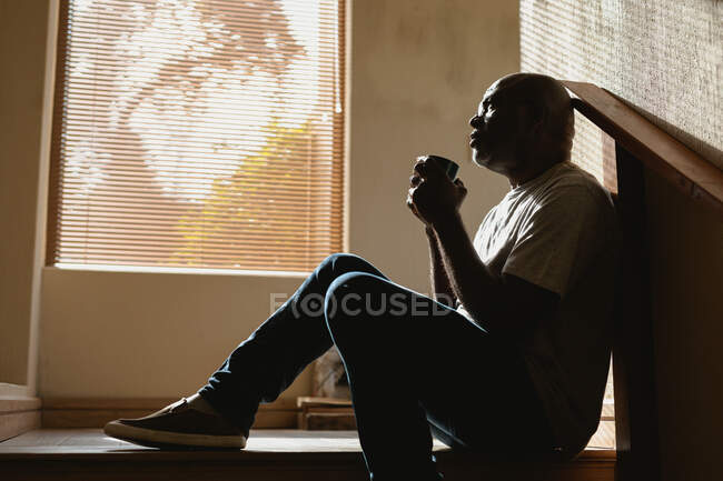 Um pensativo afro-americano sênior sentado nas escadas e bebendo café. estilo de vida aposentadoria, passar o tempo sozinho em casa. — Fotografia de Stock