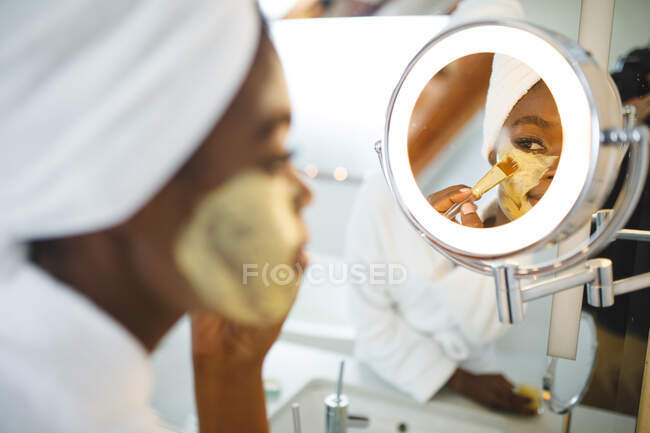 Smiling african american woman in bathroom applying beauty face mask, looking in mirror. domestic lifestyle, enjoying self care leisure time at home. — Stock Photo