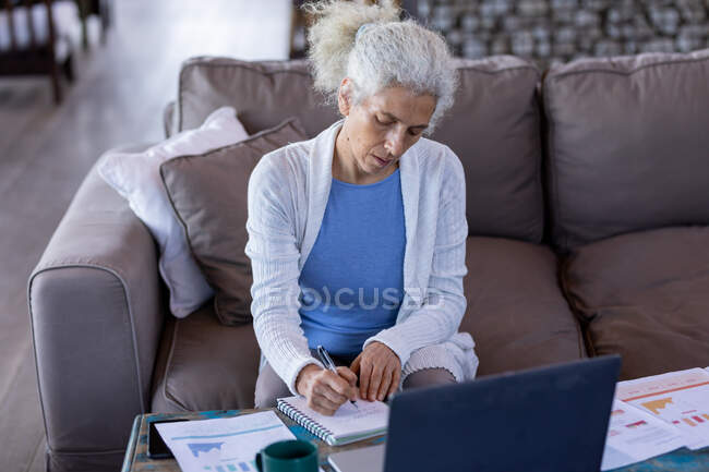 Eine ältere kaukasische Frau im Wohnzimmer sitzt auf der Couch und macht sich Notizen. Lebensstil im Ruhestand, Zeit allein zu Hause verbringen. — Stockfoto