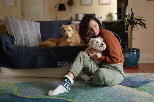 Улыбающаяся белая женщина в гостиной, сидящая на полу и обнимающая свою собаку. домашний образ жизни, наслаждаясь отдыхом дома. — стоковое фото