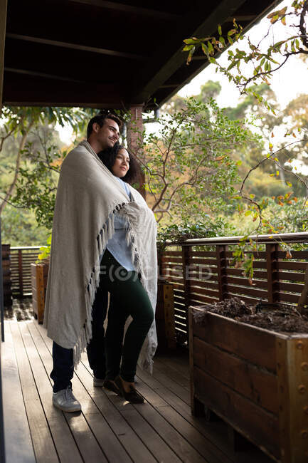 Щаслива різноманітна пара вкрита ковдрою на балконі, що охоплює і посміхається. проводити вихідні вдома . — стокове фото