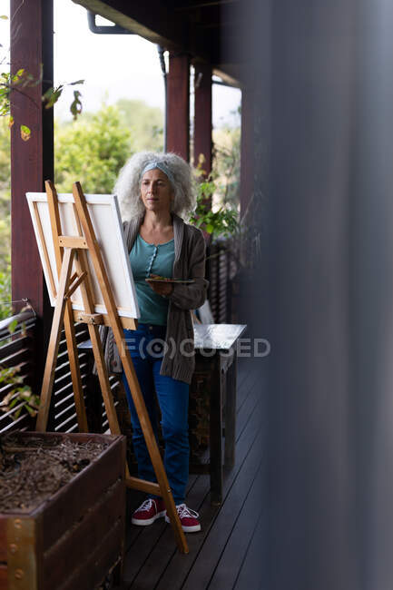 Eine ältere kaukasische Frau steht auf einem sonnigen Balkon und malt. Lebensstil im Ruhestand, Zeit allein zu Hause verbringen. — Stockfoto