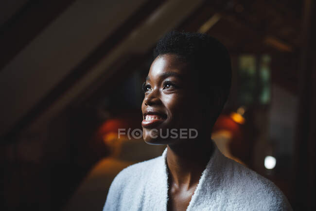 Smiling african american woman in bathroom wearing bathrobe, looking away. domestic lifestyle, enjoying self care leisure time at home. — Stock Photo