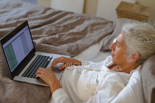 Ältere kaukasische Frau im Schlafzimmer, im Bett sitzend und mit Laptop. Lebensstil im Ruhestand, Zeit allein zu Hause verbringen. — Stockfoto
