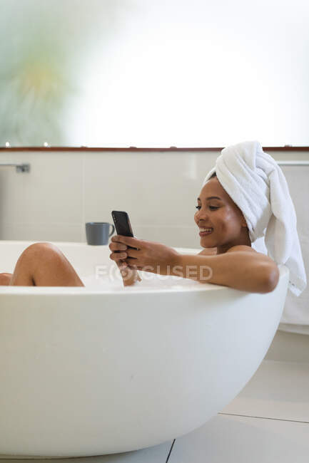 Smiling mixed race woman in bathroom having a bath and using smartphone. domestic lifestyle, enjoying self care leisure time at home. — Stock Photo