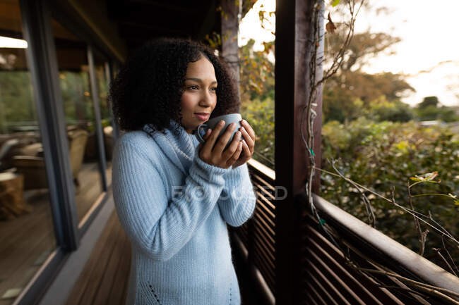 Happy mixed race woman standing on balcony holding mug and smiling. spending time off at home. — Stock Photo